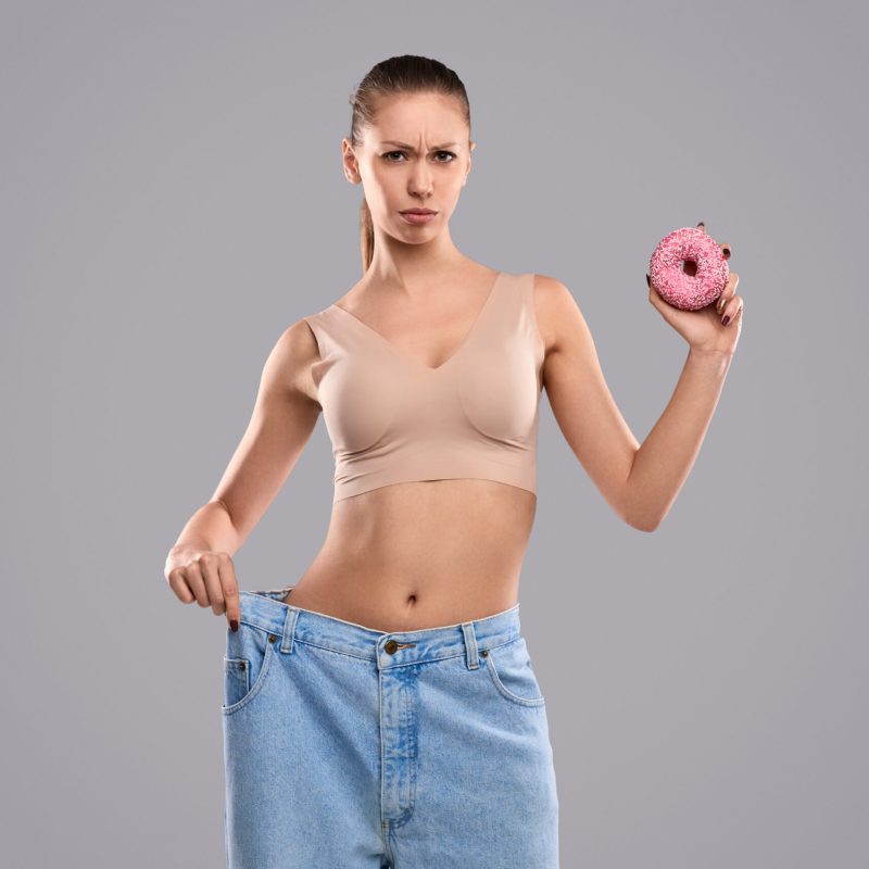 Frowning young lady in oversized jeans struggling between weight loss and unhealthy food against gray background